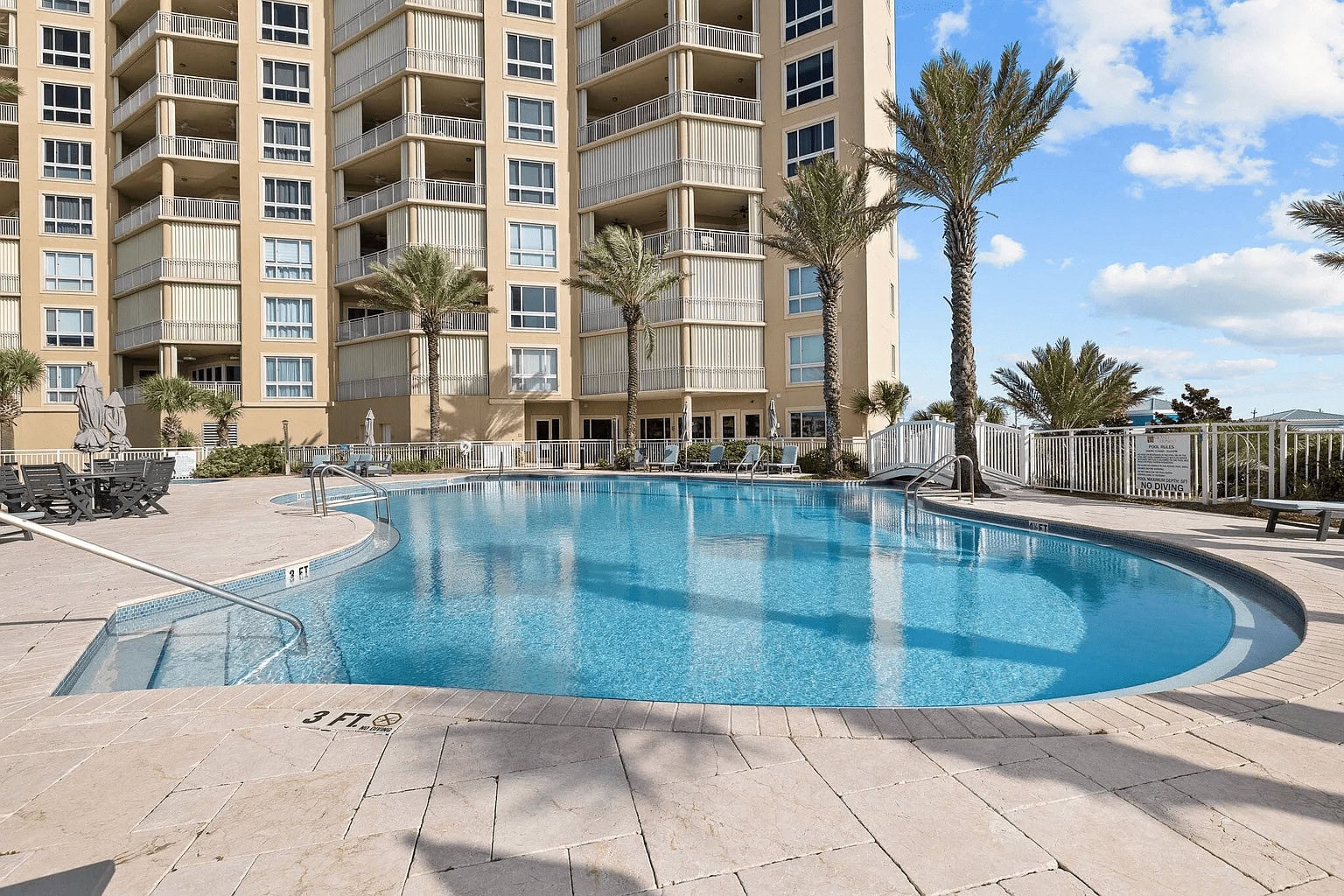 Pool area with clear blue water surrounded by a building and palm trees.